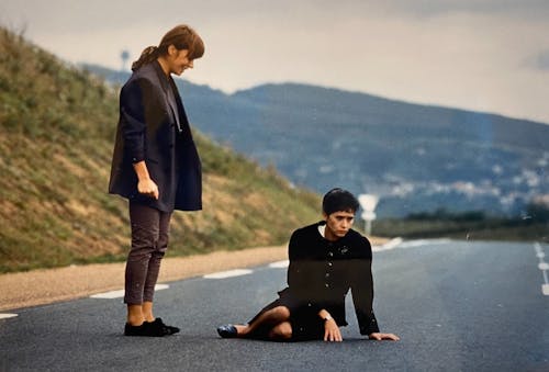 Two women on a road in a hilly area. One woman stands looking at the other who is sitting in the middle of the road looking down.