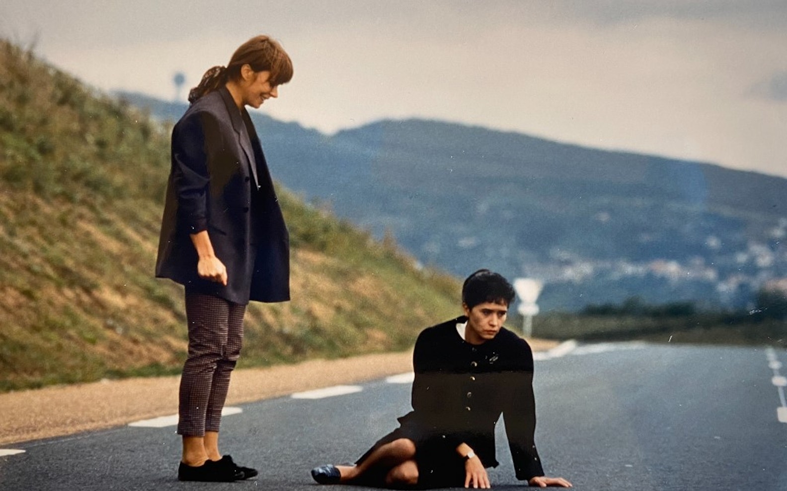 Charef_Au_pays_des_Juliets_01 Two women on a road in a hilly area. One woman stands looking at the other who is sitting in the middle of the road looking down.