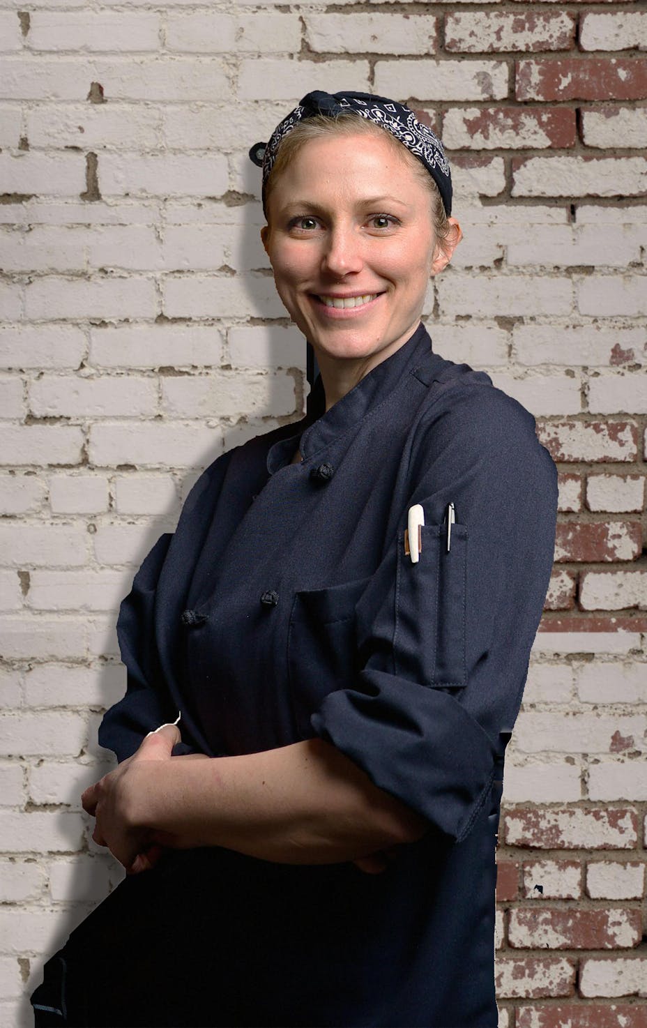 A smiling woman in a black chef's coat and bandana stands with crossed arms in front of a white brick wall. She has a pen and a thermometer tucked into her sleeve pocket.