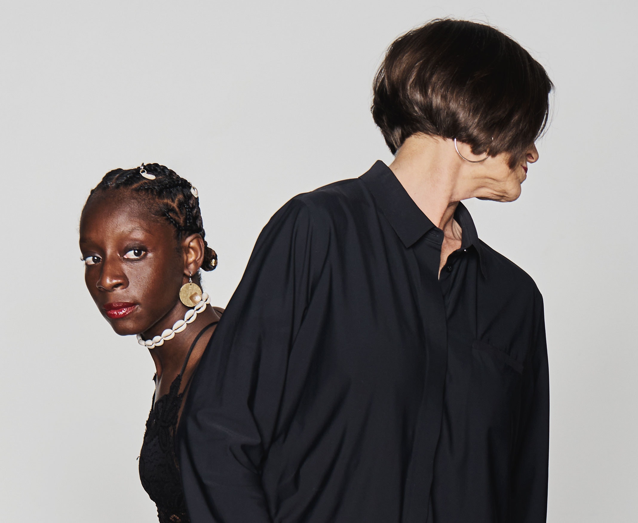 Two women stand next to one another, with one turned away from the viewer and the other looking around at the viewer.