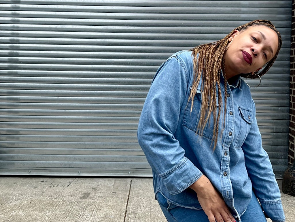 An adult with medium dark skin and long light brown braids, wearing all denim, smiles and look at the viewer mid pose.