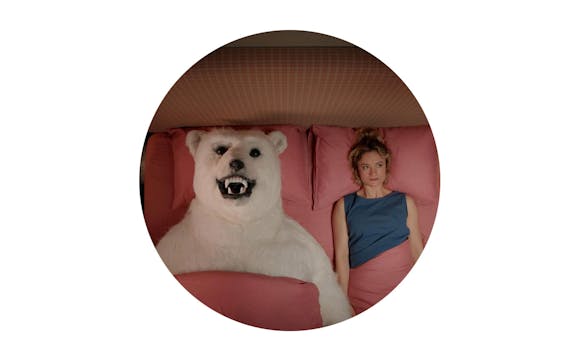 A woman with light skin lays next to a polar bear in a bed.