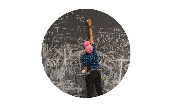 A young person wearing a baseball cap jumps up to draw on a wall using chalk.