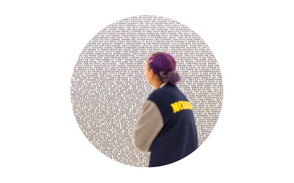 A person with purple hair pulled back wearing a varsity jacket looks at a large painitng of black words in lines.