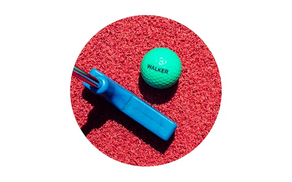 A green mini golf ball with the word "Walker" on it sits on red astroturf with a mini gulf club next to it.