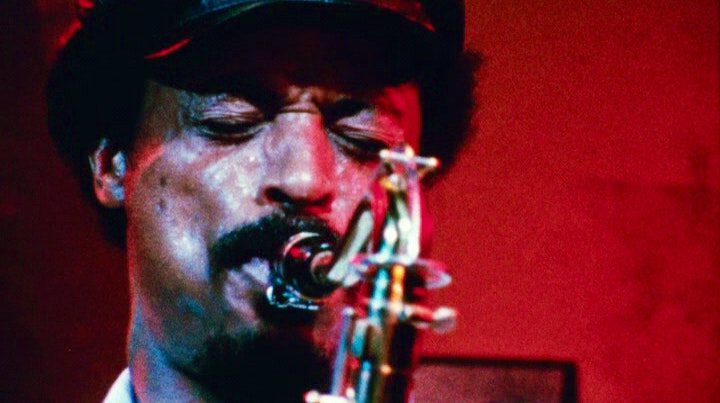 Very close still of a man playing a saxophone with his eyes squeezed shut, surrounded by red light.