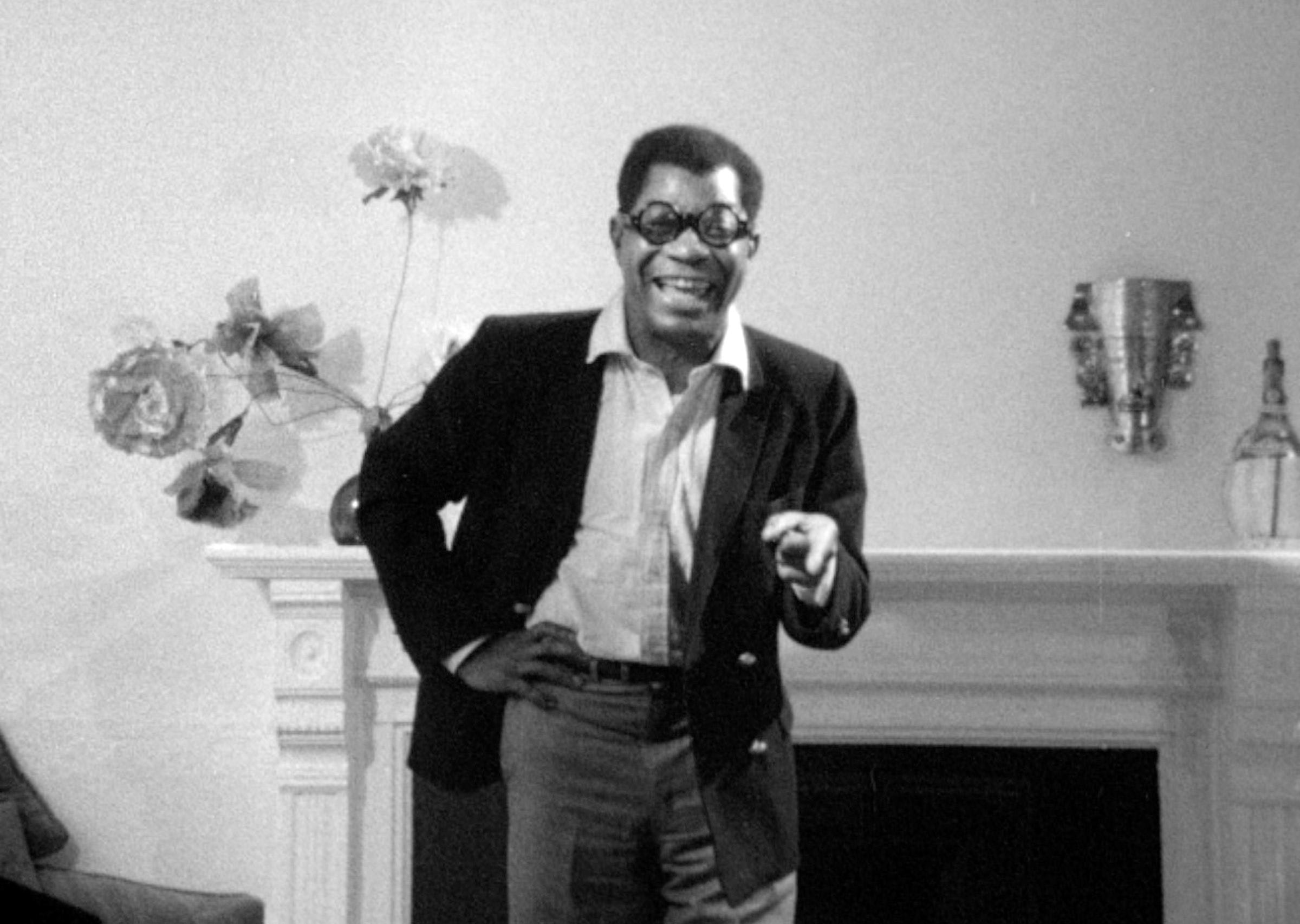 Black and white still of a man in front of a fireplace in a sport coat and round glasses, laughing and pointing at the camera.