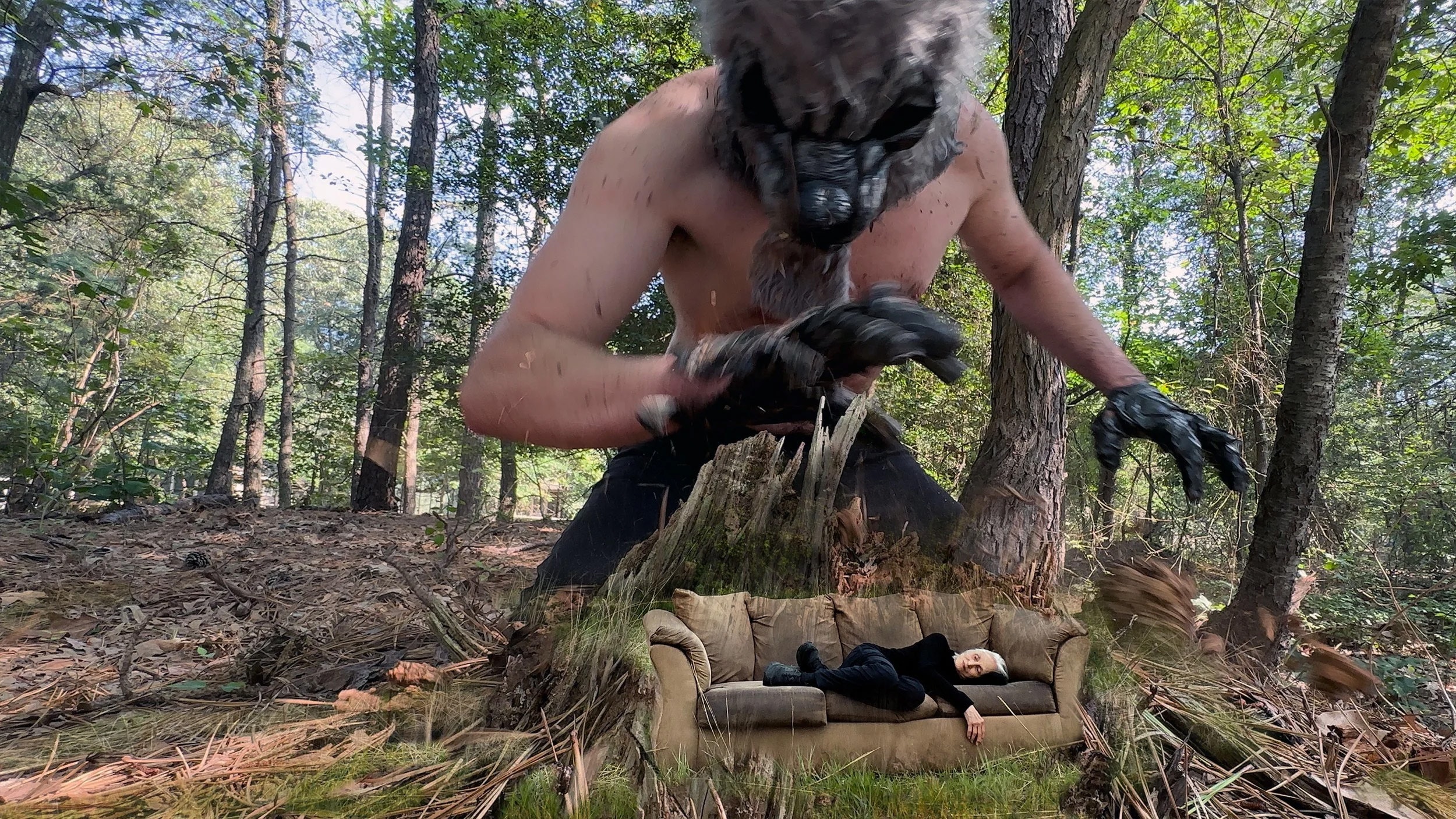 A woman in black clothing lays on a brown couch in the woods, composited with a man in the gloves and mask of a bear creature, towering over her.
