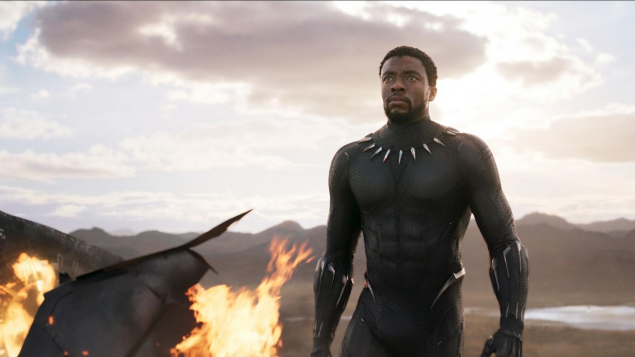 A man with dark skin wearing a black superhero suit up to his neck, and walking determinedly from some burning wreckage in a mountainous landscape.