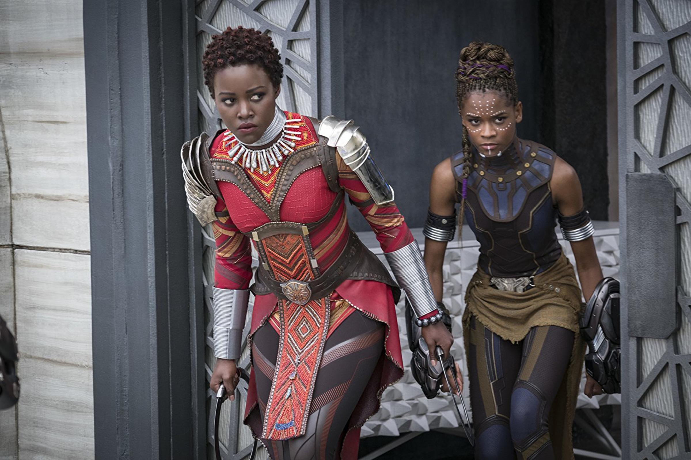 Two women with dark skin lean cautiously out of a doorway with weapons in hand. They wear decorative clothing , jewelry and hairstyles.