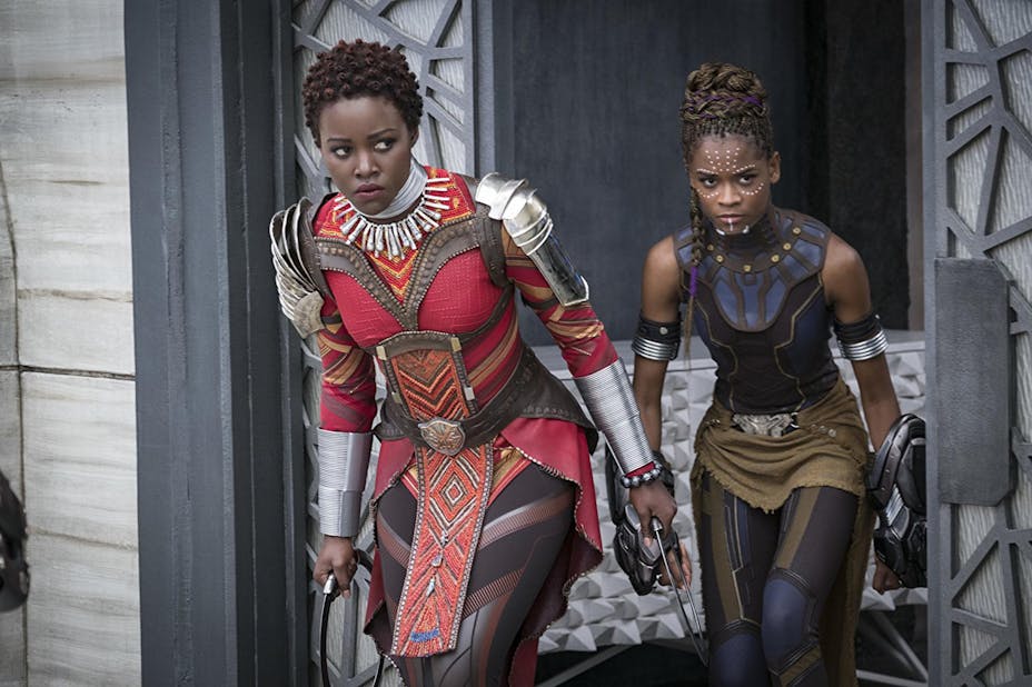 Two women with dark skin lean cautiously out of a doorway with weapons in hand. They wear decorative clothing , jewelry and hairstyles.