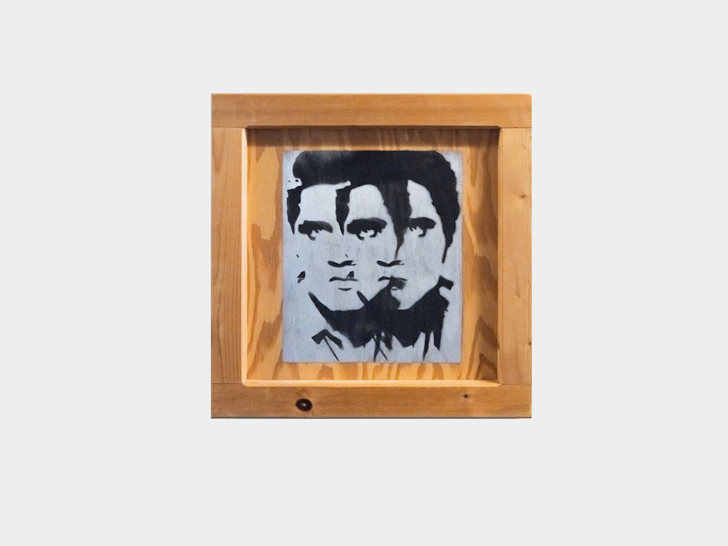 A square light wood piece of art with two black stencil faces overlapping on a metallic background.
