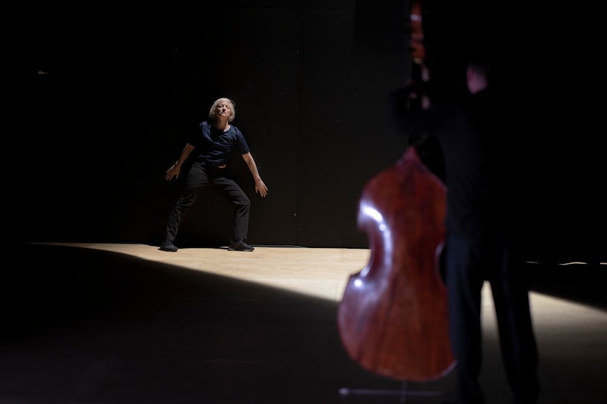 A light-skinned woman stands against a black wall facing a person with a cello.