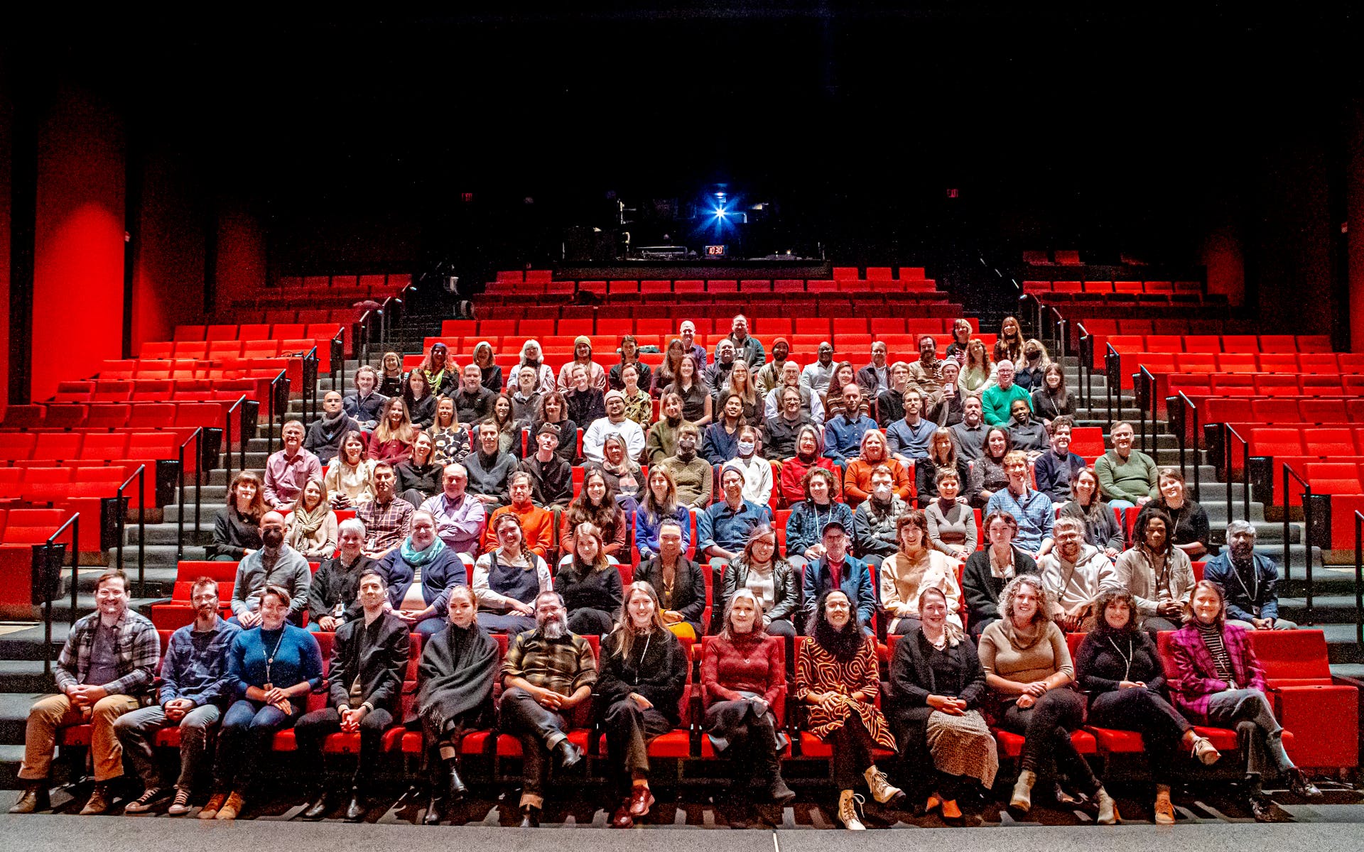A group of adults sit in a theater audience looking at the viewer and smiling.