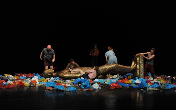 A group of actors movve around an overturned statue and piles of plastic bags on a stage.