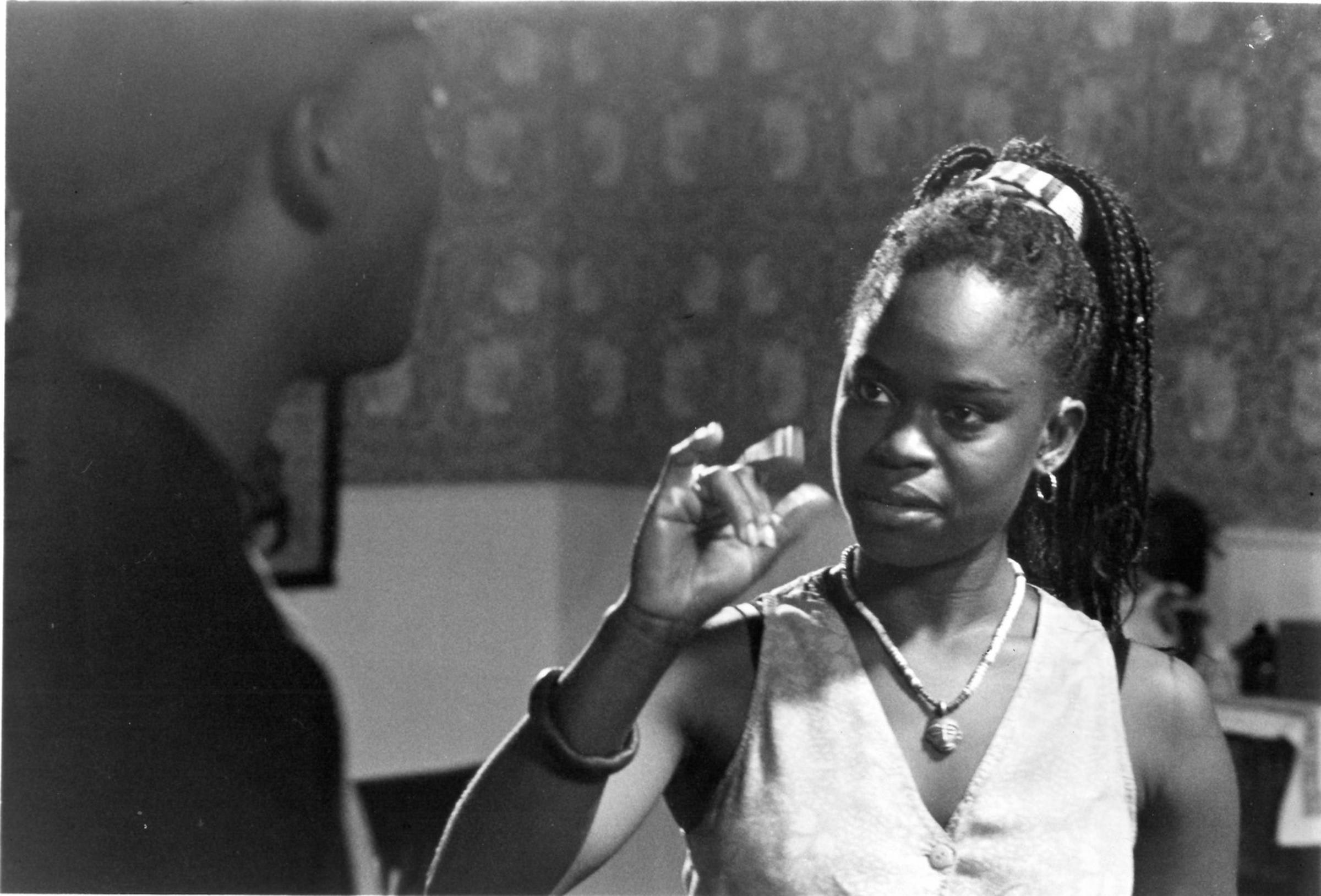 A black and white still of a Black woman wearing a vest and beaded necklace signing ASL to a Black man in the foreground.