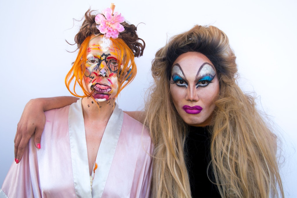 An adult wearing a black ball-cap applies make-up to another adult.