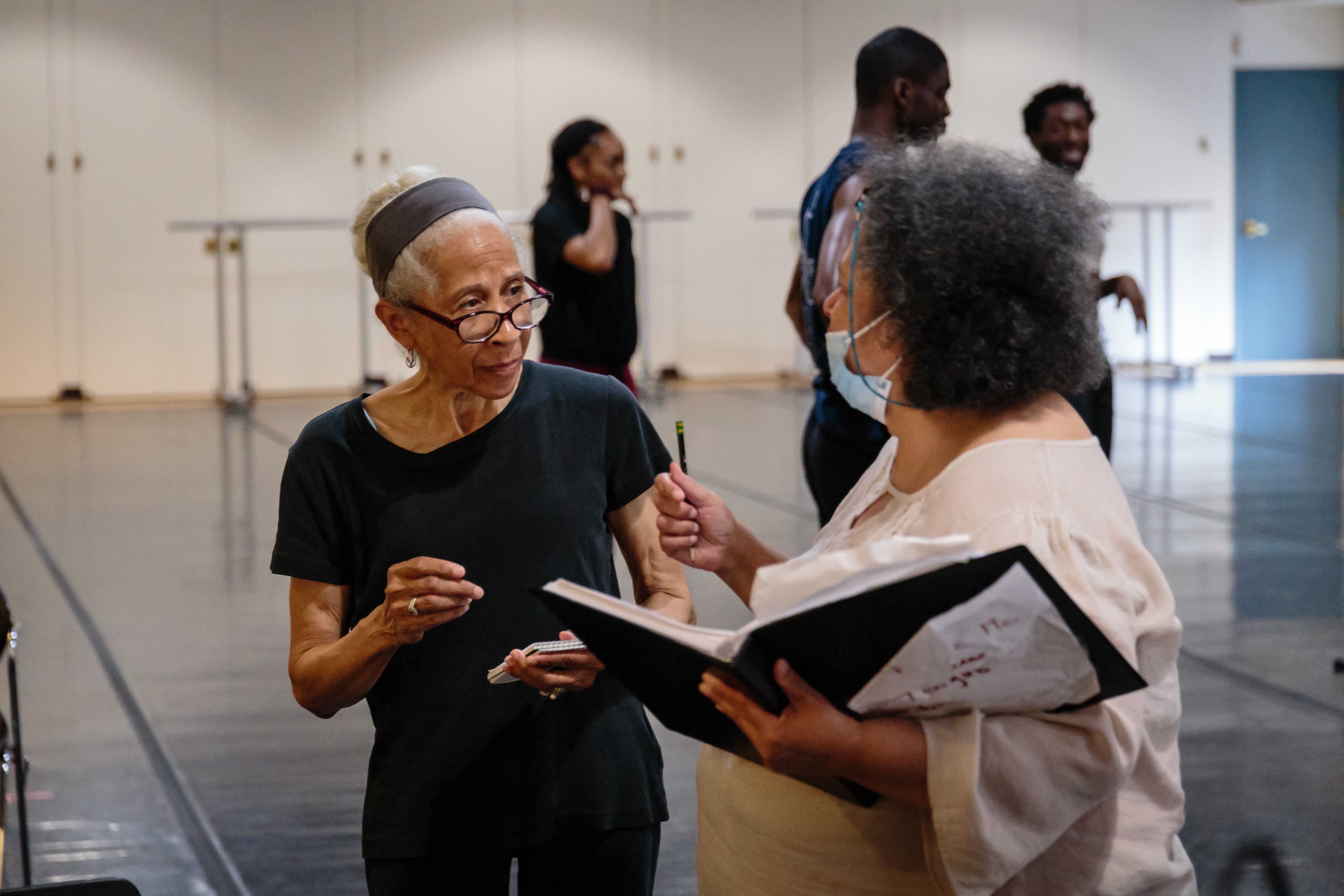 A woman with dark skin wearing glasses speaks to another woman with dark skin wearing a face mask inf ront of a group of three dancers with dark skin in a dance studio.