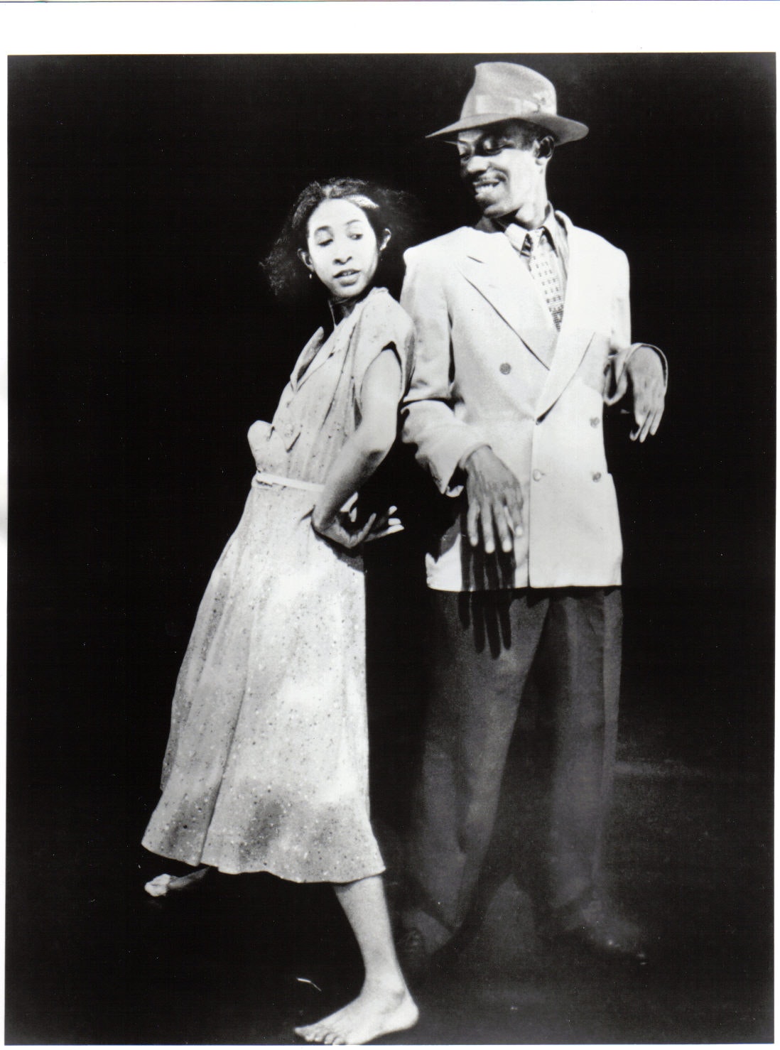 A dancer in a dress leans up against another dance in a full suit and hat on stage.