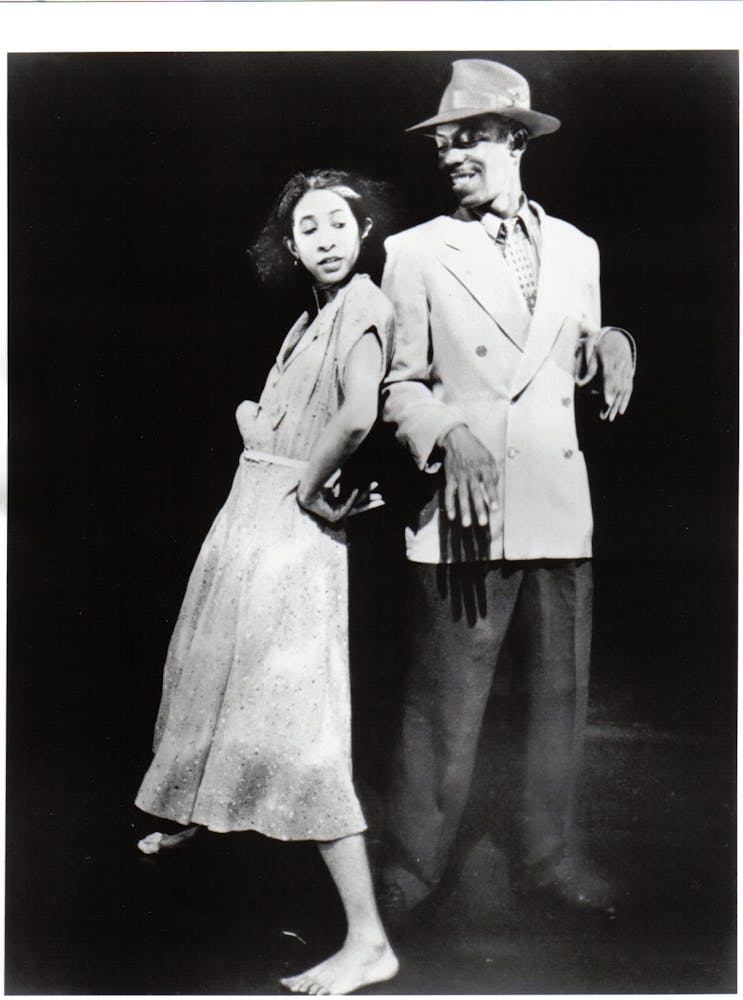 A dancer in a dress leans up against another dance in a full suit and hat on stage.