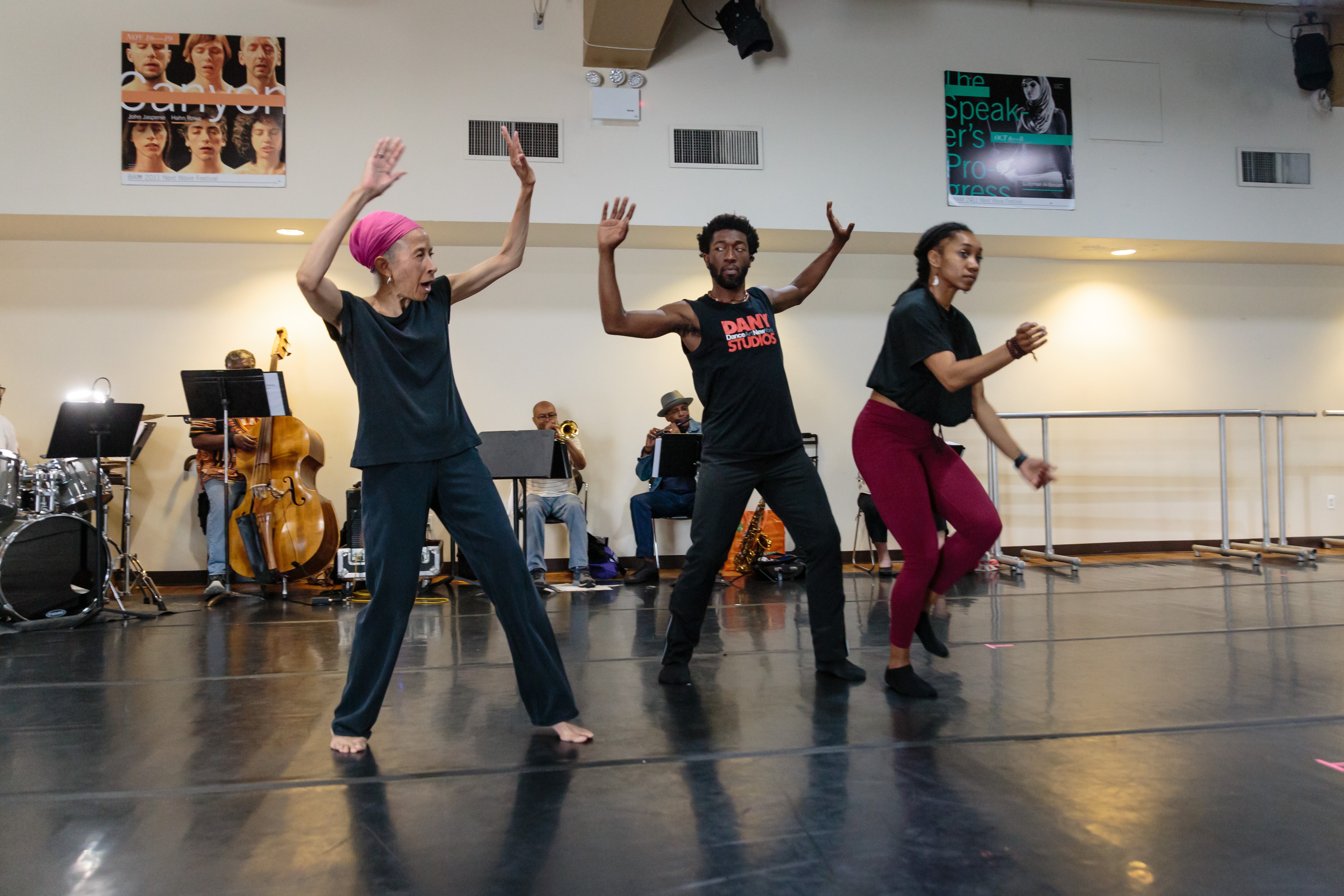 A dancer with dark skin shows two other dancers with dark skin moves in front of a band in a dance space.