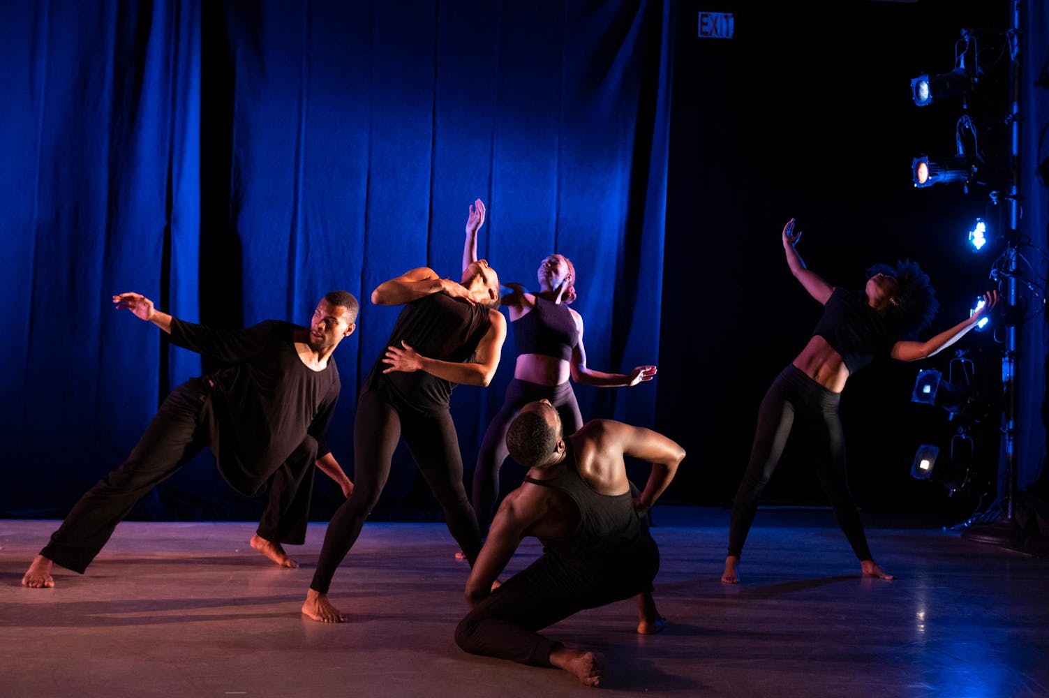 Five dancers on a stage with blue lighting seen in the background. All the dancers are wearing all black and are in various poses.