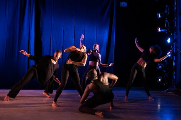 Five dancers on a stage with blue lighting seen in the background. All the dancers are wearing all black and are in various poses.