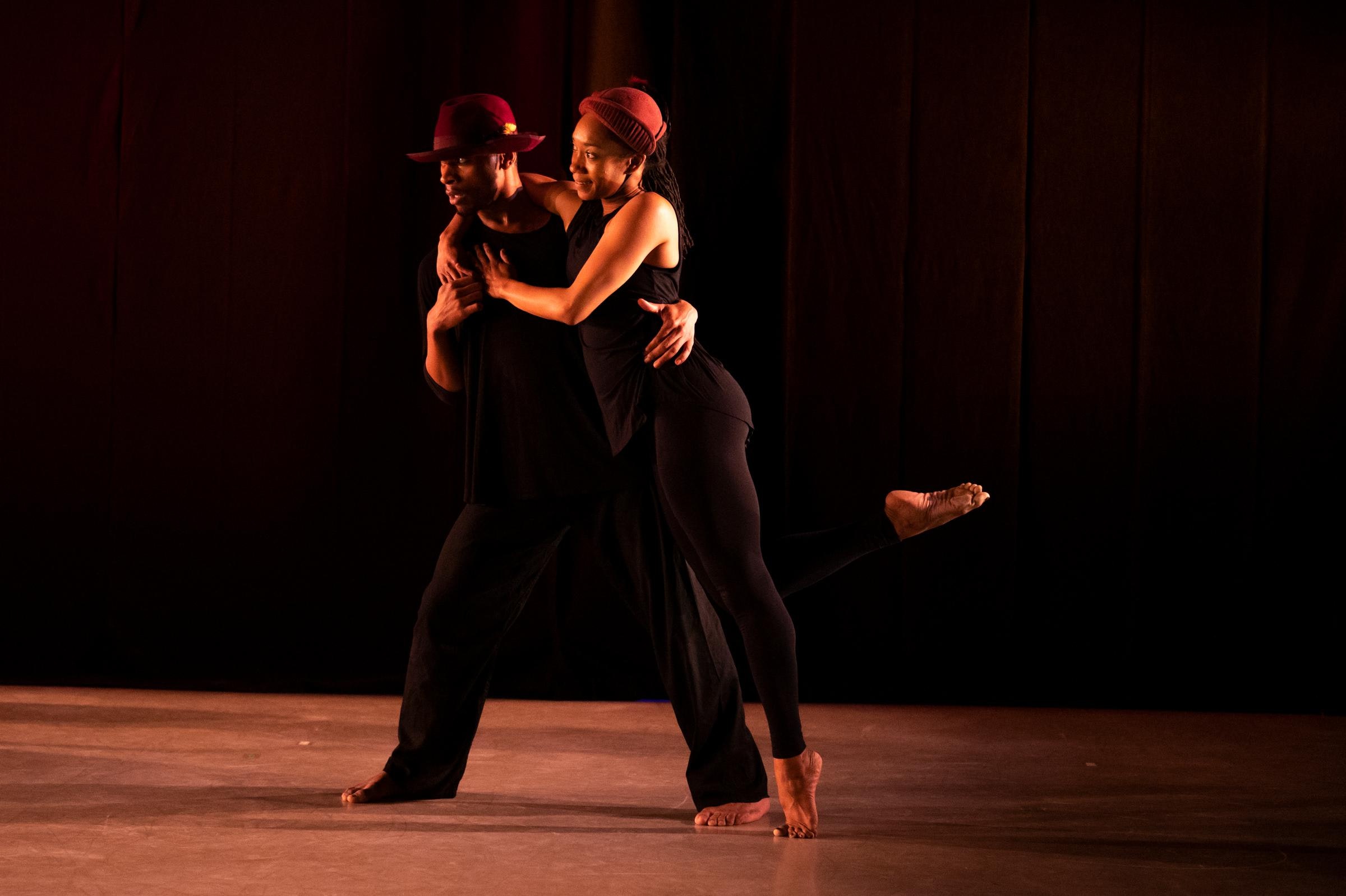 Two dancers, both wearing all black and both with dark skin and different red hats on, hold each other while standing on stage. One has their right foot pointed behind them.