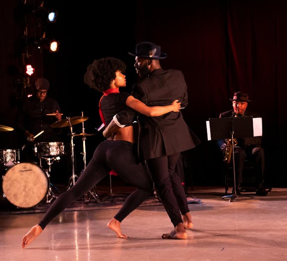 Two dancers with dark skin, one wearing a suit and hat and the other wearing a red scar, tango on a stage with two jazz musicians seen behind them.