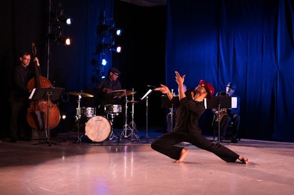 A dancer onstage with dark skin and a red hat, with legs bent low to the ground and left leg stretched far behind them, and index and middle fingers pointed in front of them. Four jazz musicians are in the background.