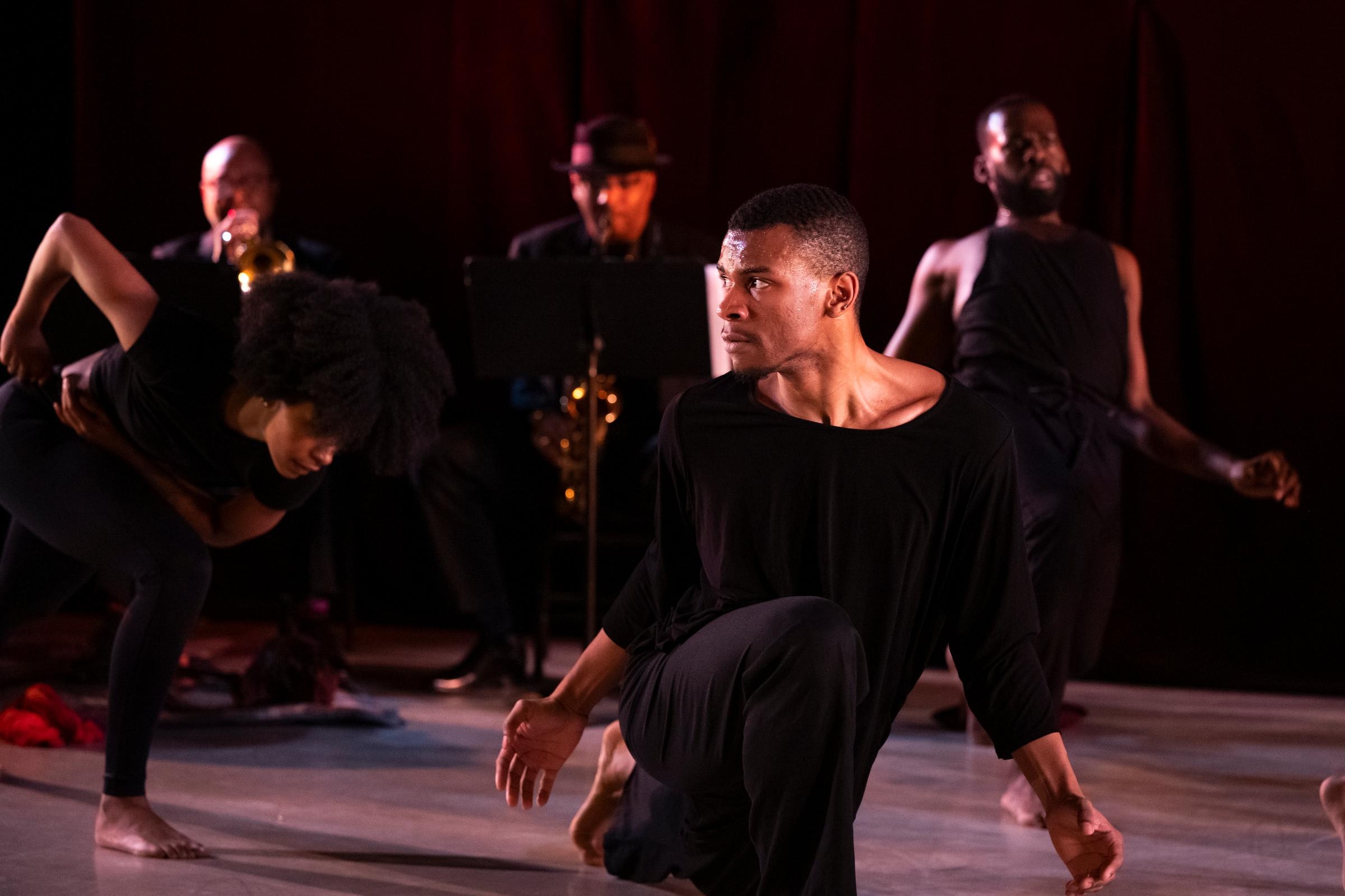 A dancer in front is on one knee with their hands beside them. In the background, two other dancers in various poses are seen, along with two musicians behind them.