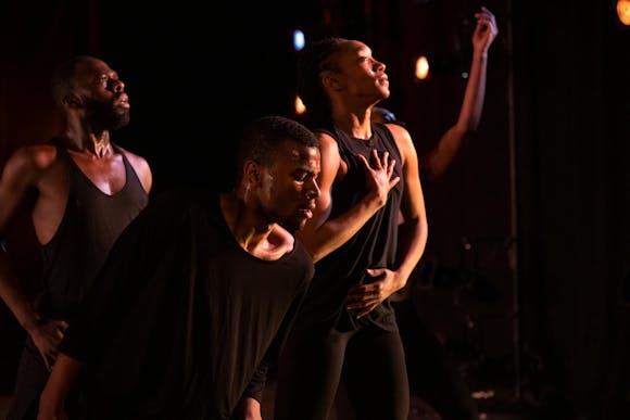 Three dancers, all with dark skin and dark hair and wearing black, face to the right. A fourth dancer is partially obscured in the background.