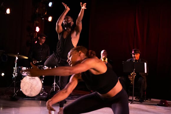 Two dancers with dark skin and dark hair, both wearing black, pose with their arms in front of them on a stage. Three musicians, a drummer, trumpeter, and saxophonist are seen in the background.