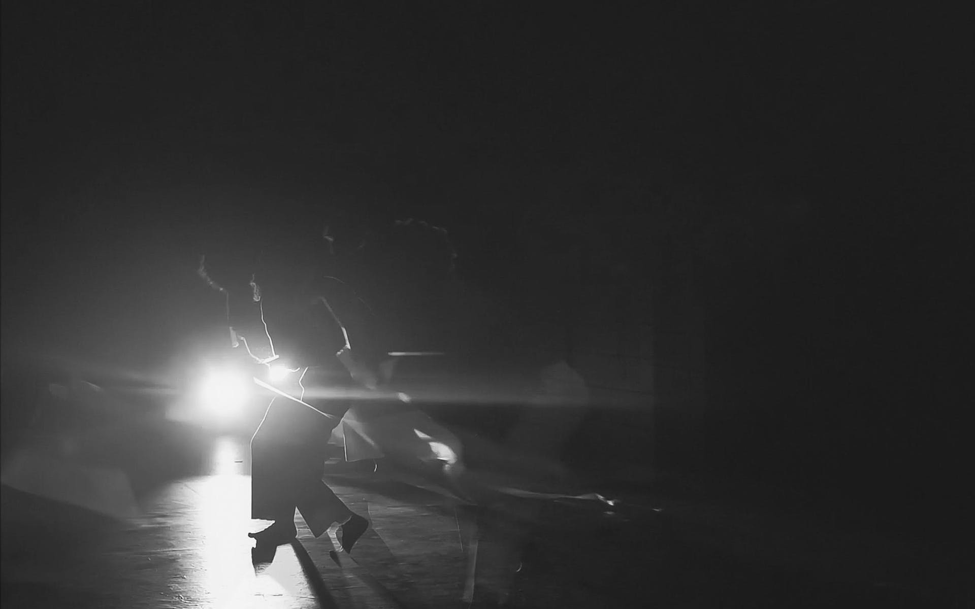 Black and white image of a person squating in front of a white light on a dark stage.