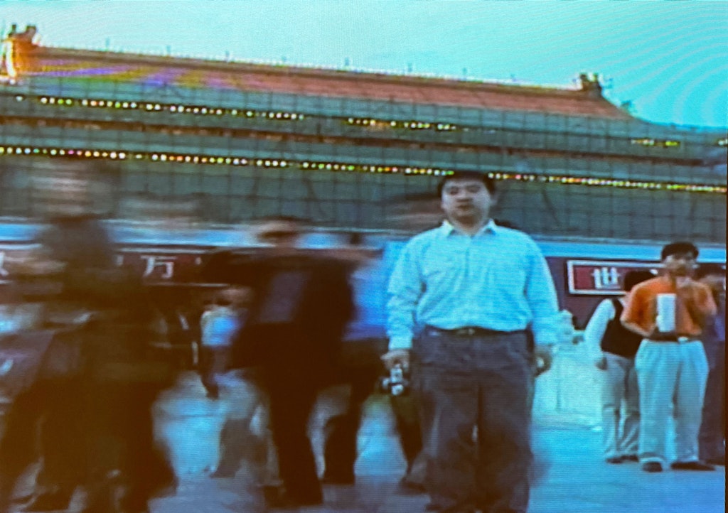 An adult wearing a white button up shirt and dark pants walks towards viewer, people blurred in photograph around them in front of a green building.