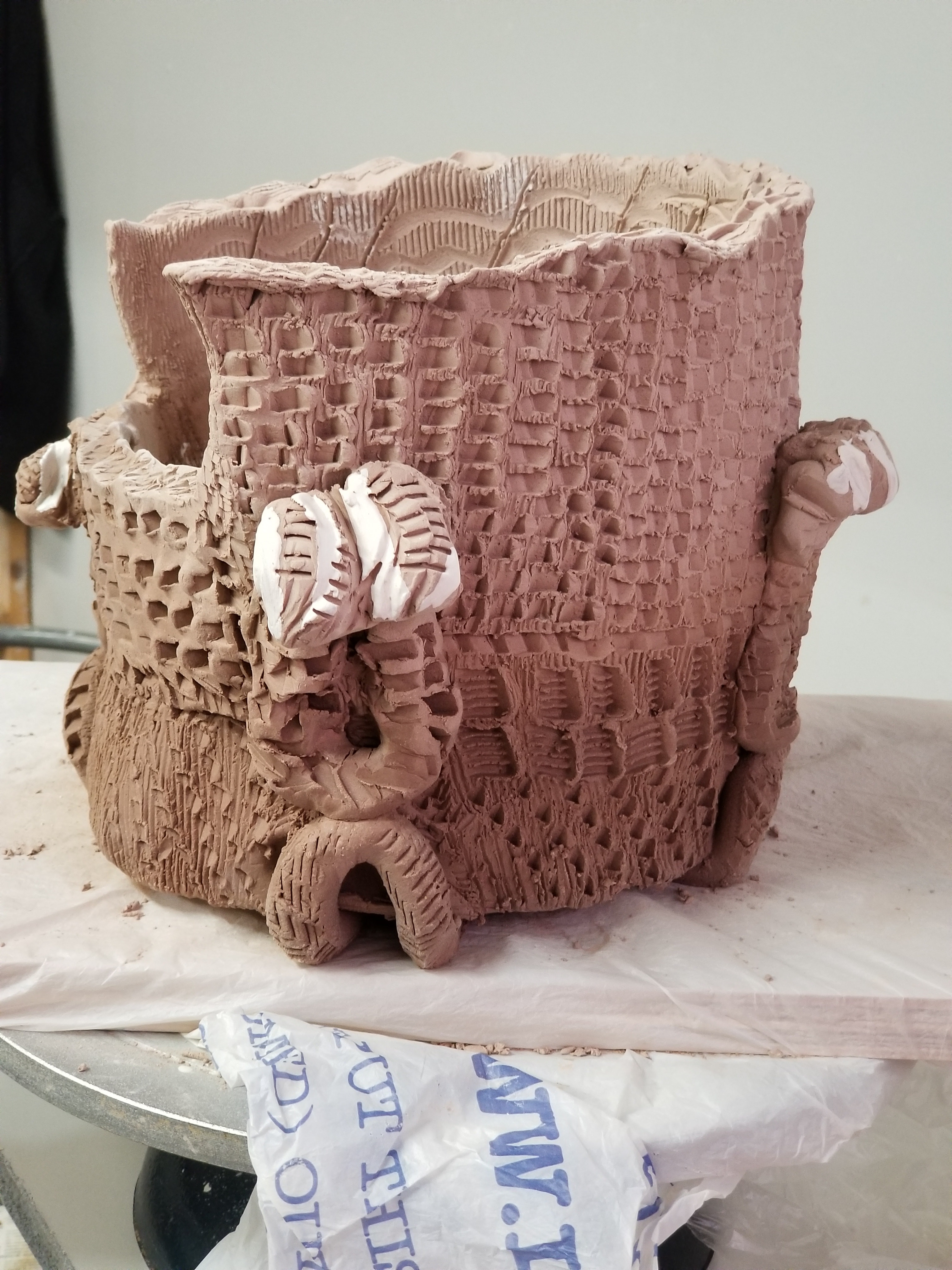 A ceramic vessel in earth-tones with an indented texture throughout. It is open to the top and has another opening on the side, and there are additional rounded pieces of clay attached at the front and sides