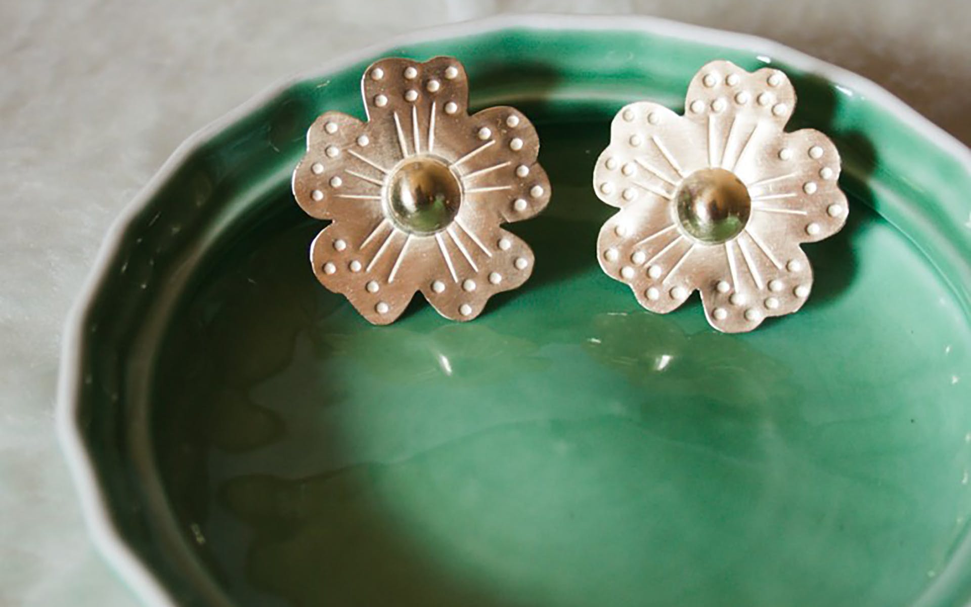 Image of flower shaped, metal earrings sitting on green ceramic dish