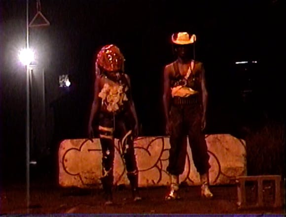Two people in distopian cowboy outfits stand in a parking lot at night.