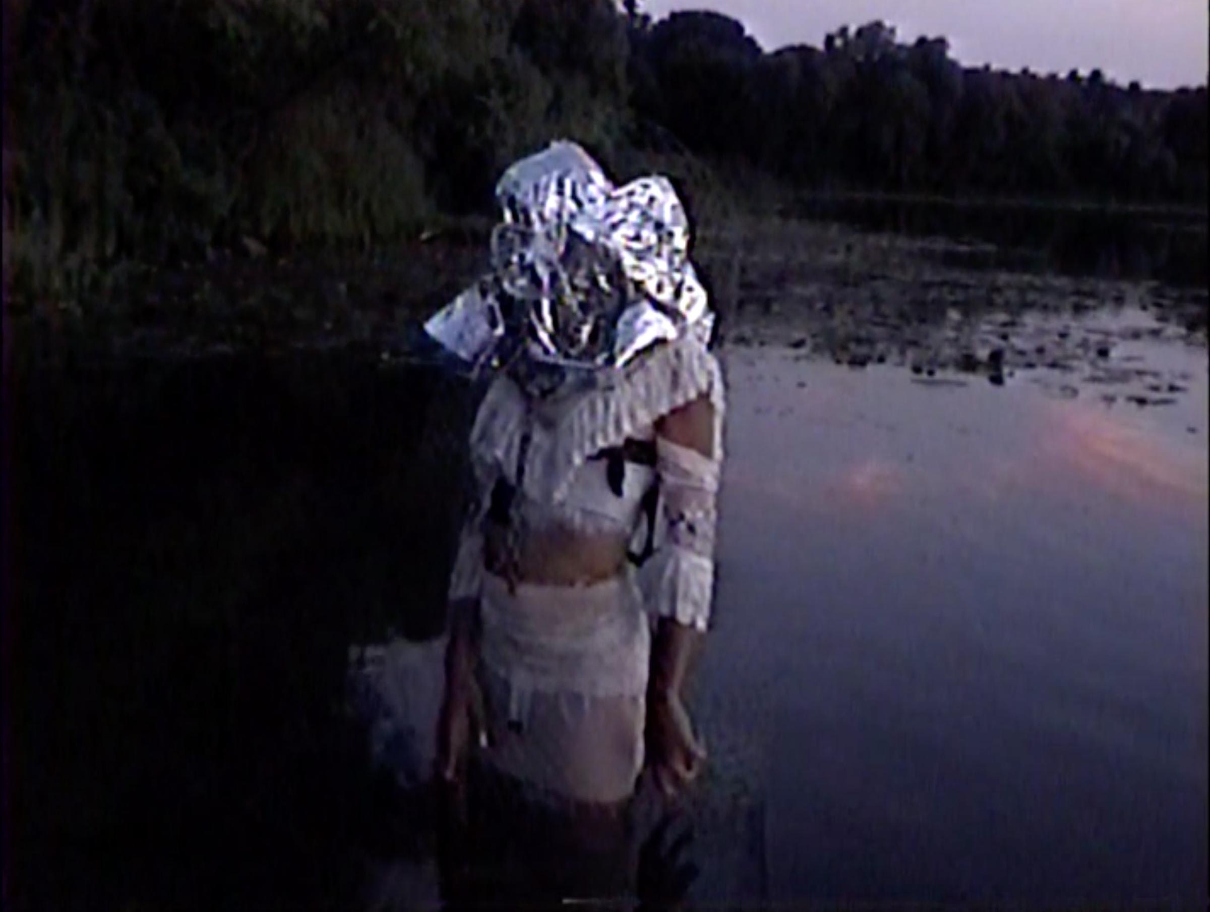 A person with silver metalic covering over their head stands in a lake.