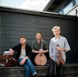 Three musicians pose with their instruments standing in front of a nondescript, grey building.