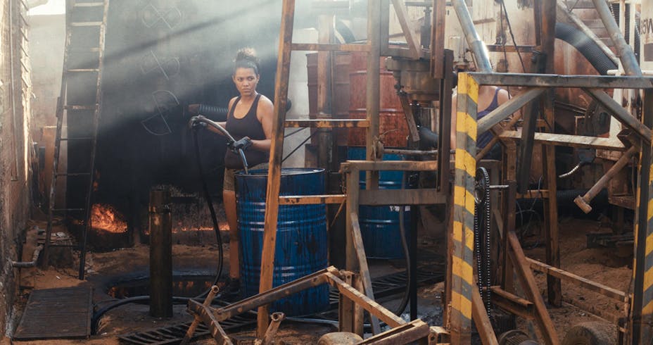 A woman stands holding a tube that leads into a metal barrel in a construction site.