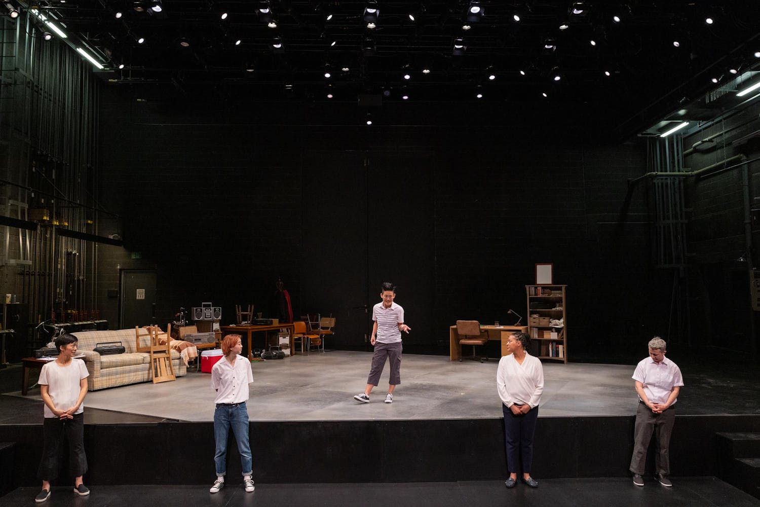Four people stand in front of a stage, while a fifth person stands on the stage, talking and gesturing with their hand. Various furniture is seen visible at the back of the stage, along with the stage lights.