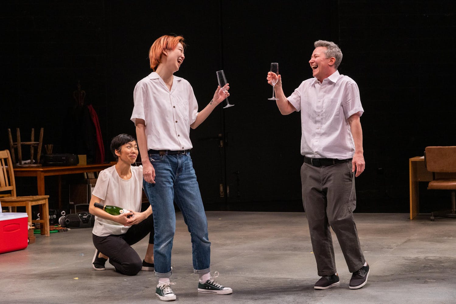 Two people with champagne glasses cheers each other and laugh while one person kneels behind them holding a bottle of champagne. Various furniture, a stereo, and a cooler are seen towards the back of the stage.