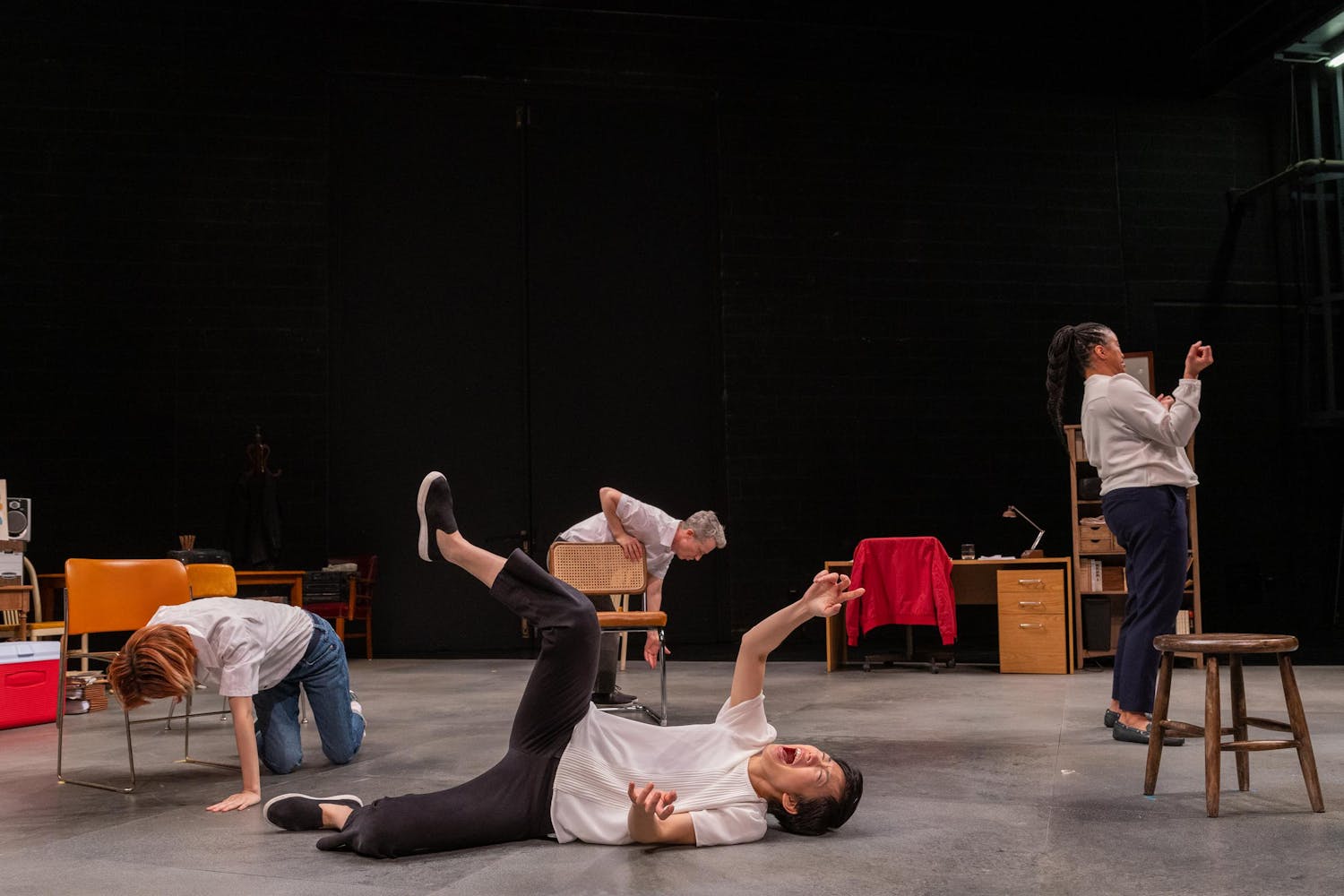 A performer with light skin and short dark hair lays on their side with one leg and one arm reaching upward; they seem to be screaming. Three other performers are seen the background along with various chairs and furniture.