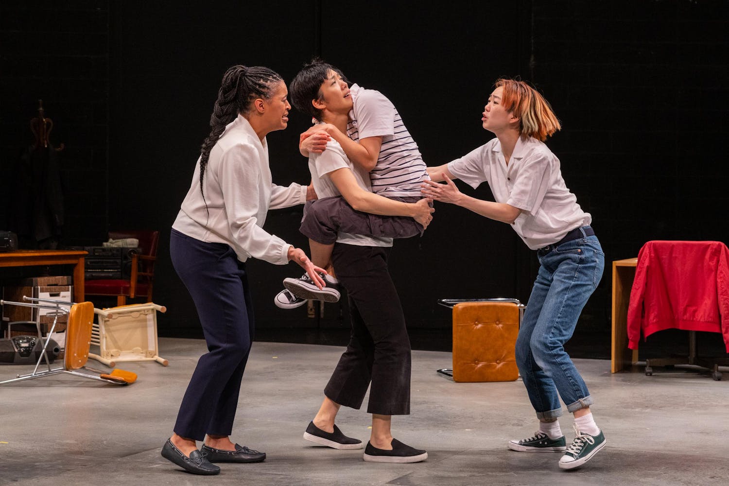 A performer stands and holds another person up, who has wrapped their legs and arms around them. Two other performers are holding their arms out, and all look concerned. Toppled furniture is seen in the background.