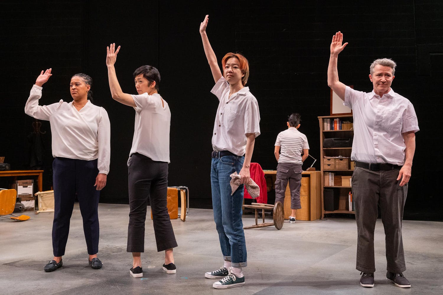 Four performers of varying skin colors and hairstyles stand at the front of a stage with their right hands raised. In the background, a figure is seen facing the back of the stage, near a desk and a bookcase.