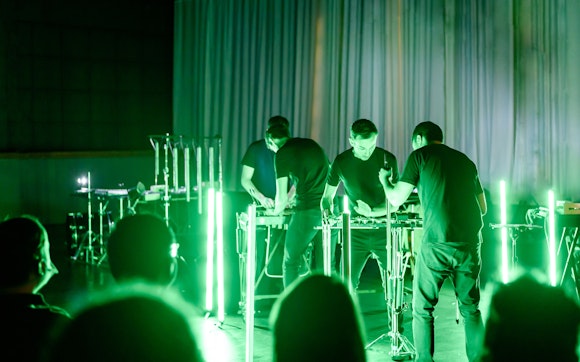 Four people with light skin and dark hair play percussion instruments on a small stage lit with green vertical fluorescent lights.