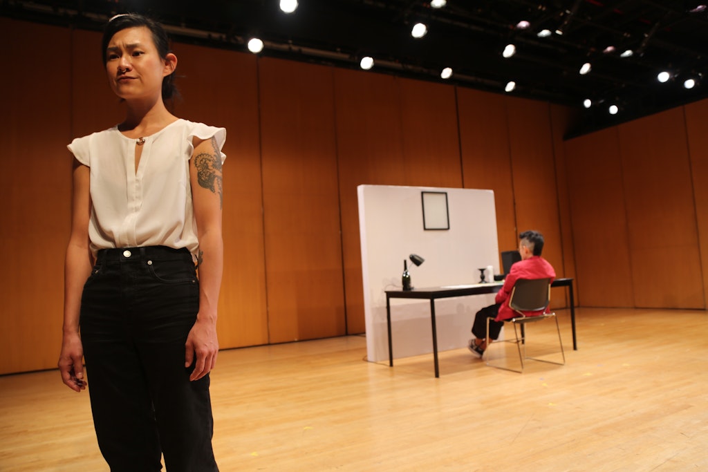 An adult wearing a white shirt and black pants on a stage stands facing the viewer in the foreground. An adult sits, facing away from viewer at a desk in background.