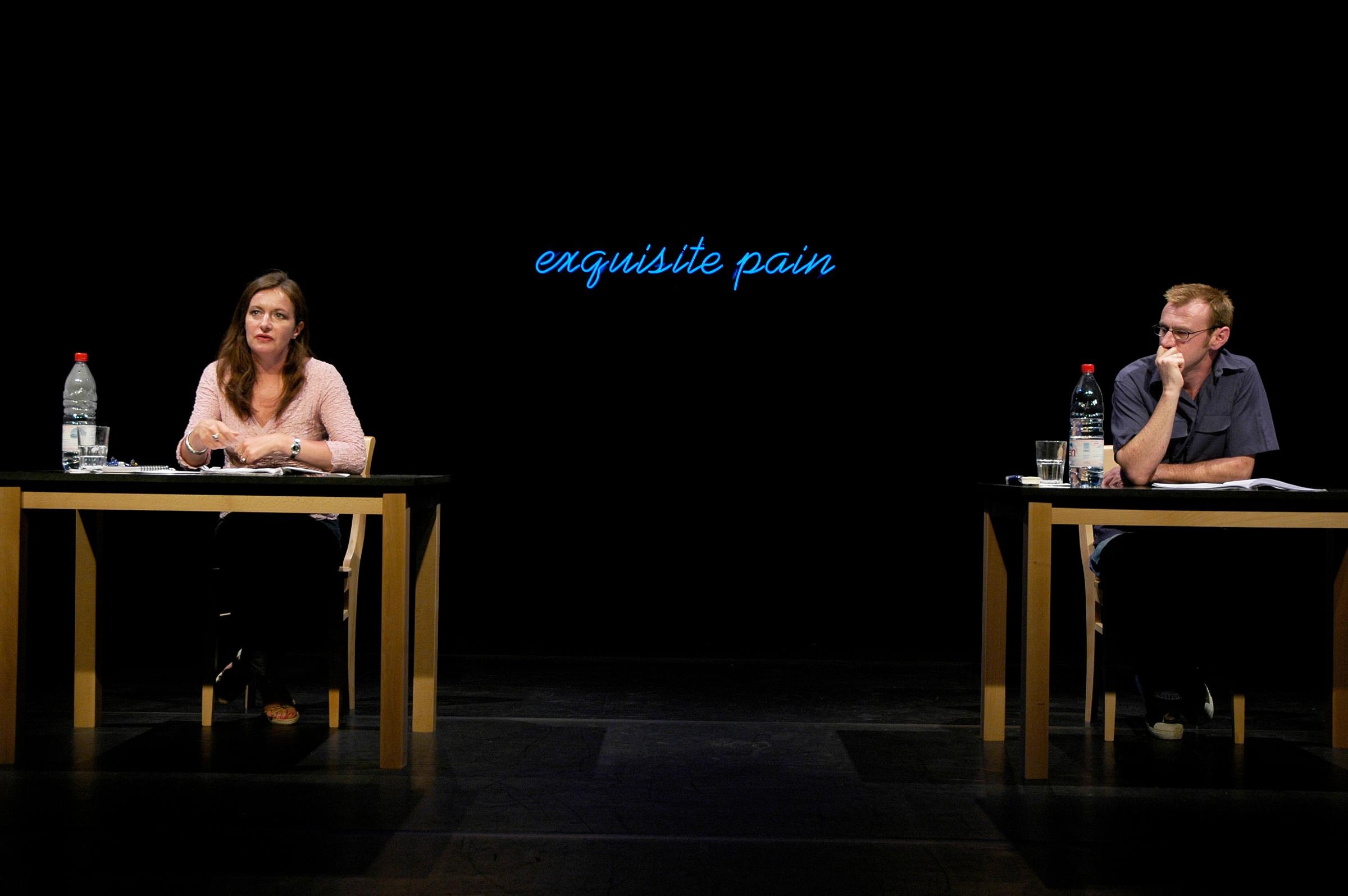 Two performers with light skin sit at matching desks in front of a neon sign that reads "exquisite pain".