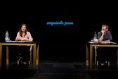 Two performers with light skin sit at matching desks in front of a neon sign that reads "exquisite pain".
